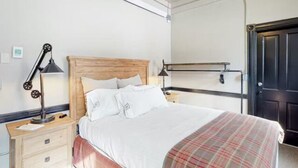 A hotel room with a wooden headboard bed, white bedding, a plaid throw blanket, a nightstand with a lamp, and a door with a black handle. - Benson Lodge Room - 6 (Silverton)