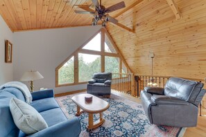 A spacious room with a high ceiling, wooden beams, a large window, a blue sofa, a grey leather chair, and a patterned rug.