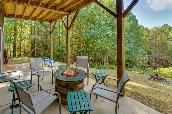 An outdoor patio with a stone fire pit surrounded by metal chairs and tables, set against a backdrop of a forested area. - Serene Cabin w/ Deck & Fire Pit in Blue Ridge Mtns (Bostic)
