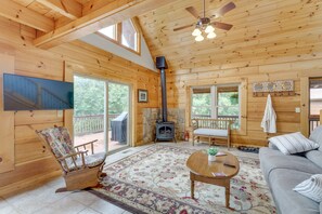 A cozy cabin interior with a fireplace, wooden walls, a ceiling fan, a sofa, a rocking chair, and a large window. - Serene Cabin w/ Deck & Fire Pit in Blue Ridge Mtns (Bostic)