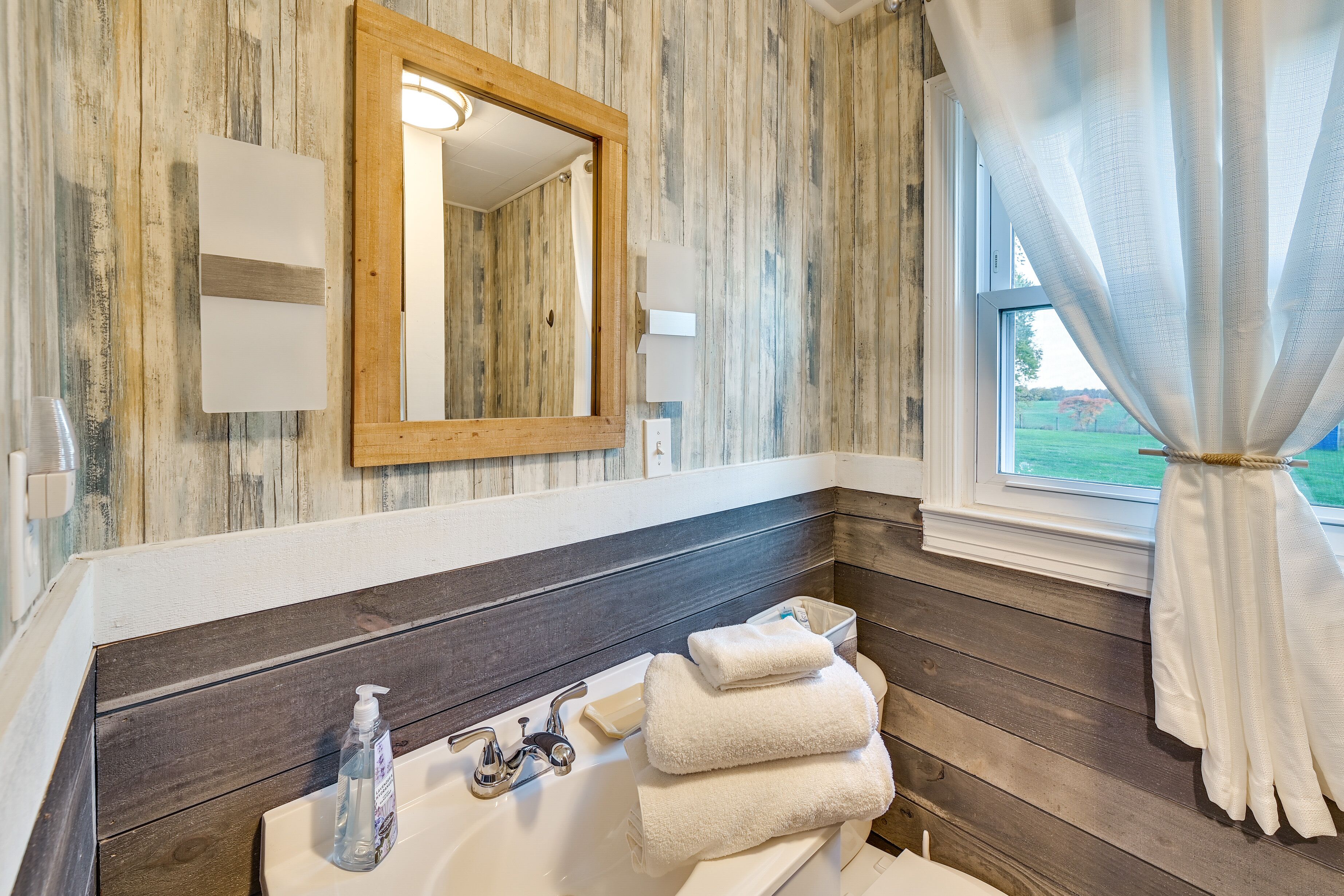 Un baño con pared de madera, un espejo, un lavabo con dispensador de jabón y una ventana con cortina.