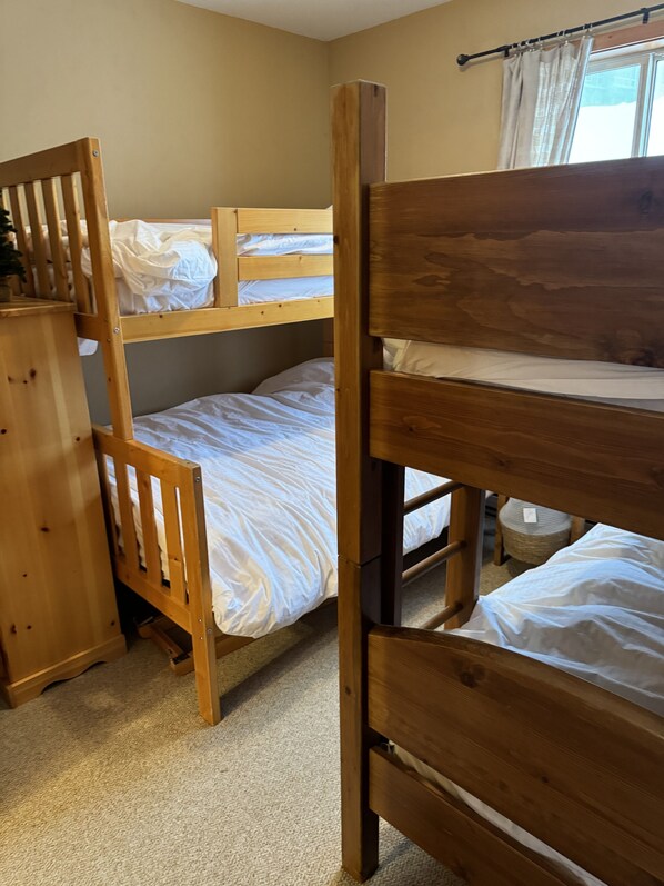 Een slaapkamer met een stapelbed, een houten hoofdbord, een klein houten dressoir en een raam met gordijnen.