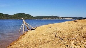 Een zandstrand met een houten constructie, kalm water en beboste heuvels op de achtergrond.