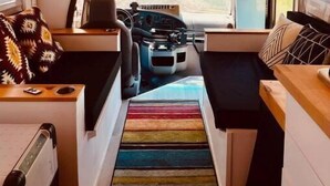 Interieur van een camper met een houten plafond, een middenbed, een kitchenette en een kleurrijk gestreept tapijt.