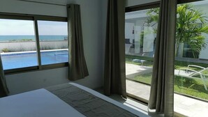 A hotel room with a bed, a window with a view of a pool and ocean, and a view of a lawn and palm trees.