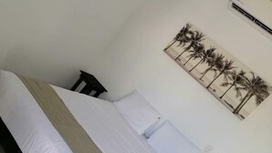A bed with white linens and a beige throw, a small bedside table, and a wall-mounted air conditioner.