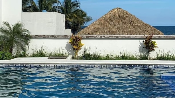 A swimming pool with a clear blue sky, a white building, and a thatched-roof structure.
