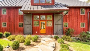 A red wooden house with a yellow door, a covered porch, and a stone pathway leading to the entrance. - The Lodge at Stoneleigh (Round Hill)