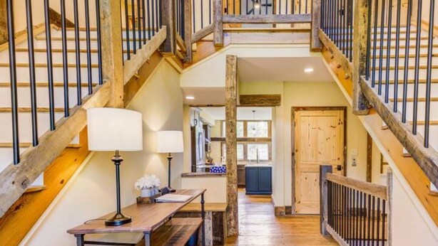 A wooden staircase with a railing, a table with a lamp, and a door in a hallway. - The Lodge at Stoneleigh (Round Hill)