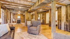 A spacious room with exposed wooden beams, a fireplace, a brown leather sofa, and a plaid armchair. - The Lodge at Stoneleigh (Round Hill)