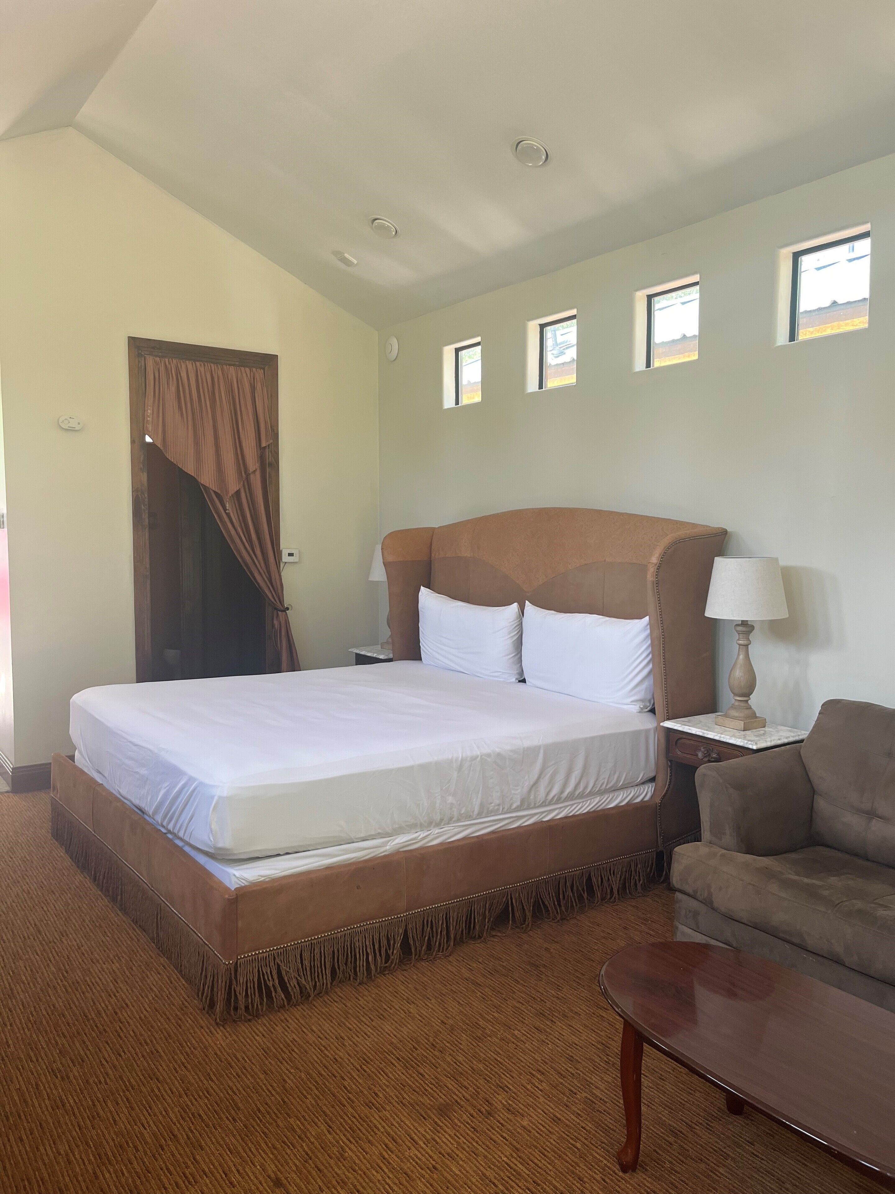 A hotel room with a large bed, a wooden headboard, a nightstand with a lamp, a small table, and a sofa.