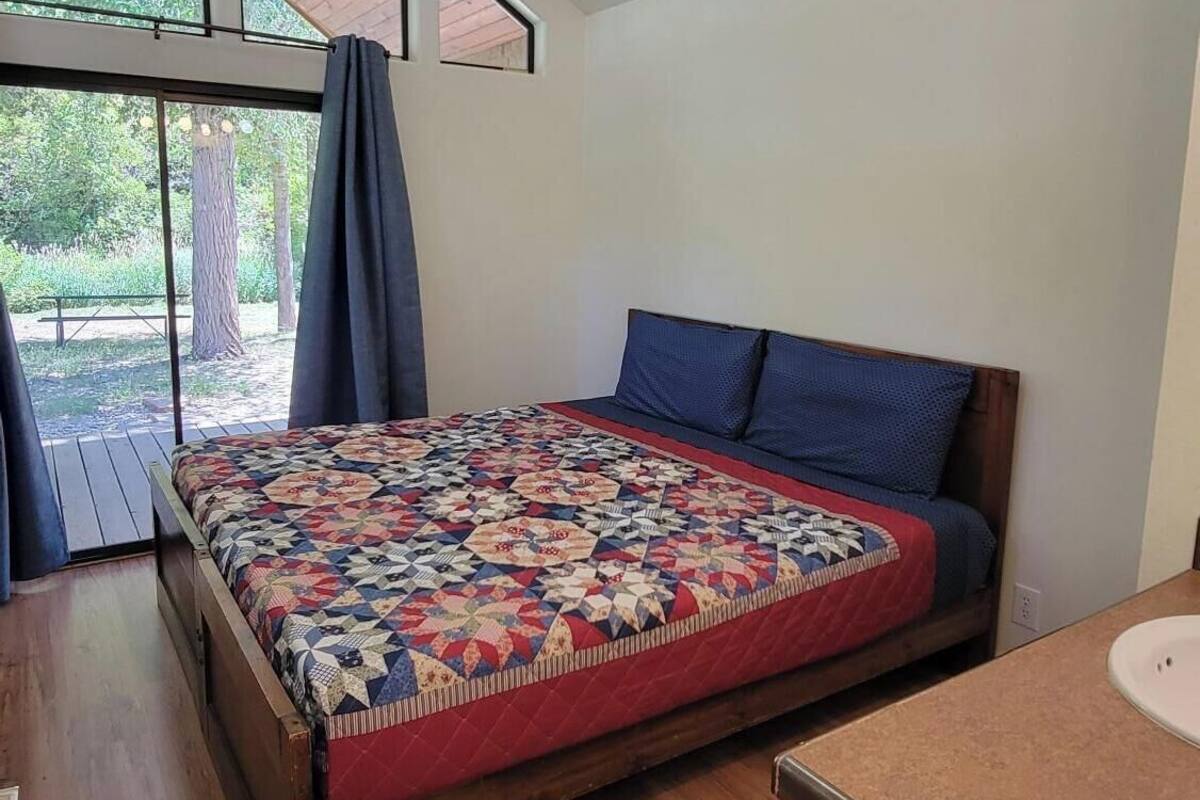 A bedroom with a wooden bed, a window with a view of greenery, and a small kitchenette area.
