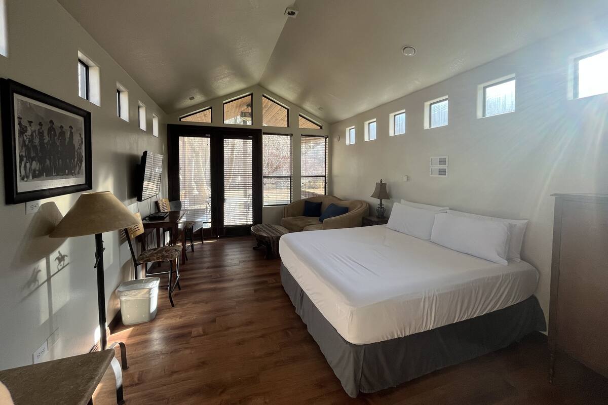 A spacious bedroom with a large bed, a desk with a chair, and a dining area. There are multiple windows providing natural light.