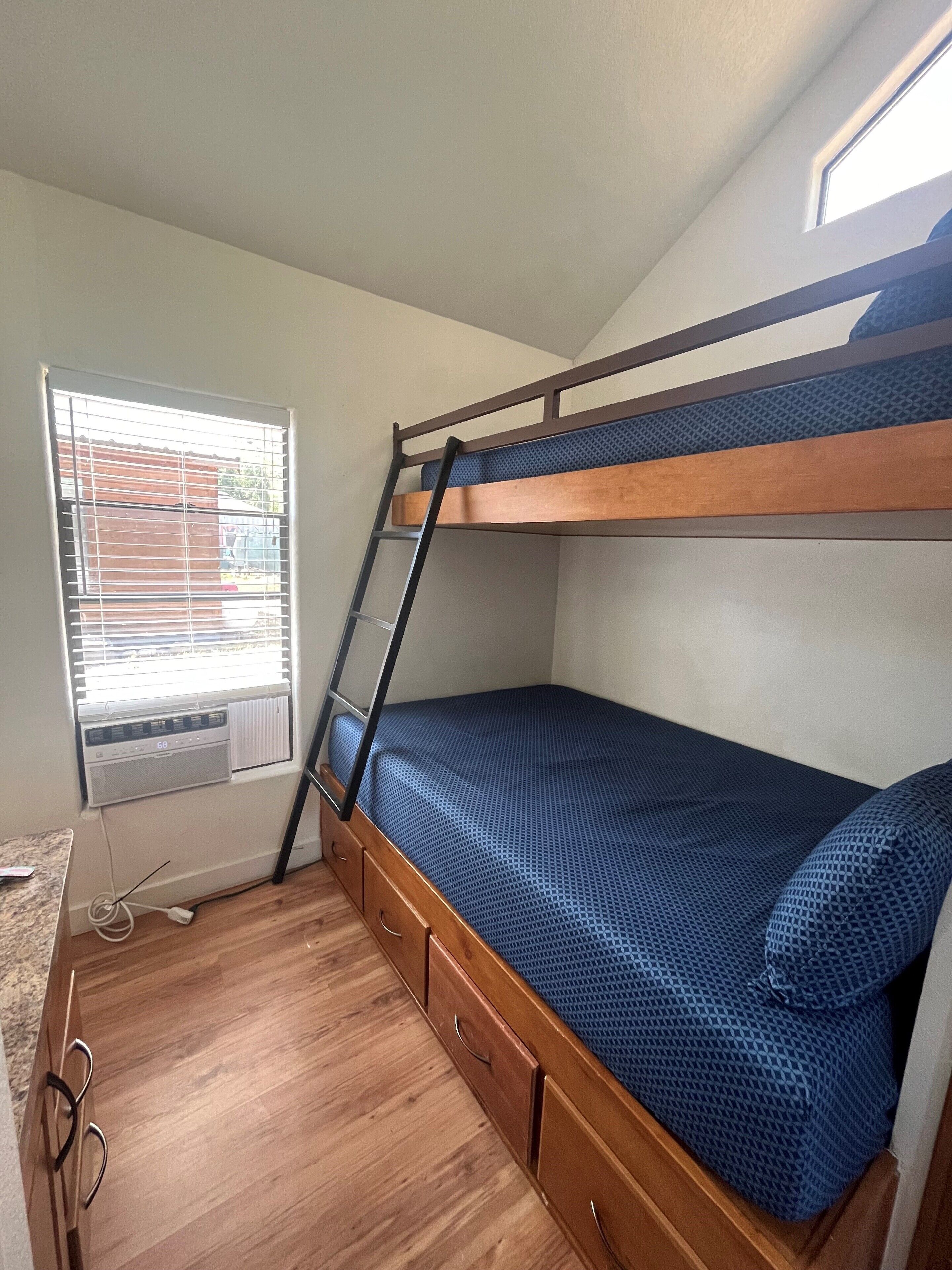 Een stapelbed met een ladder, een raam met zonwering en een houten vloer.