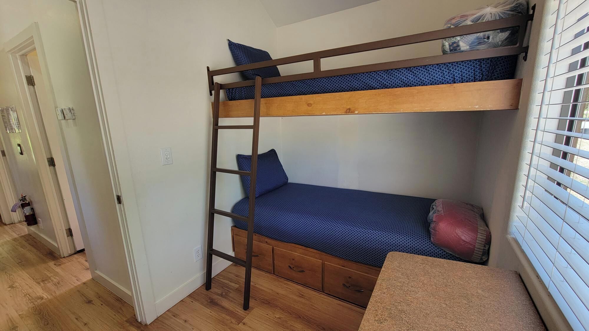 A bunk bed with a ladder, a small desk, and a window with blinds.
