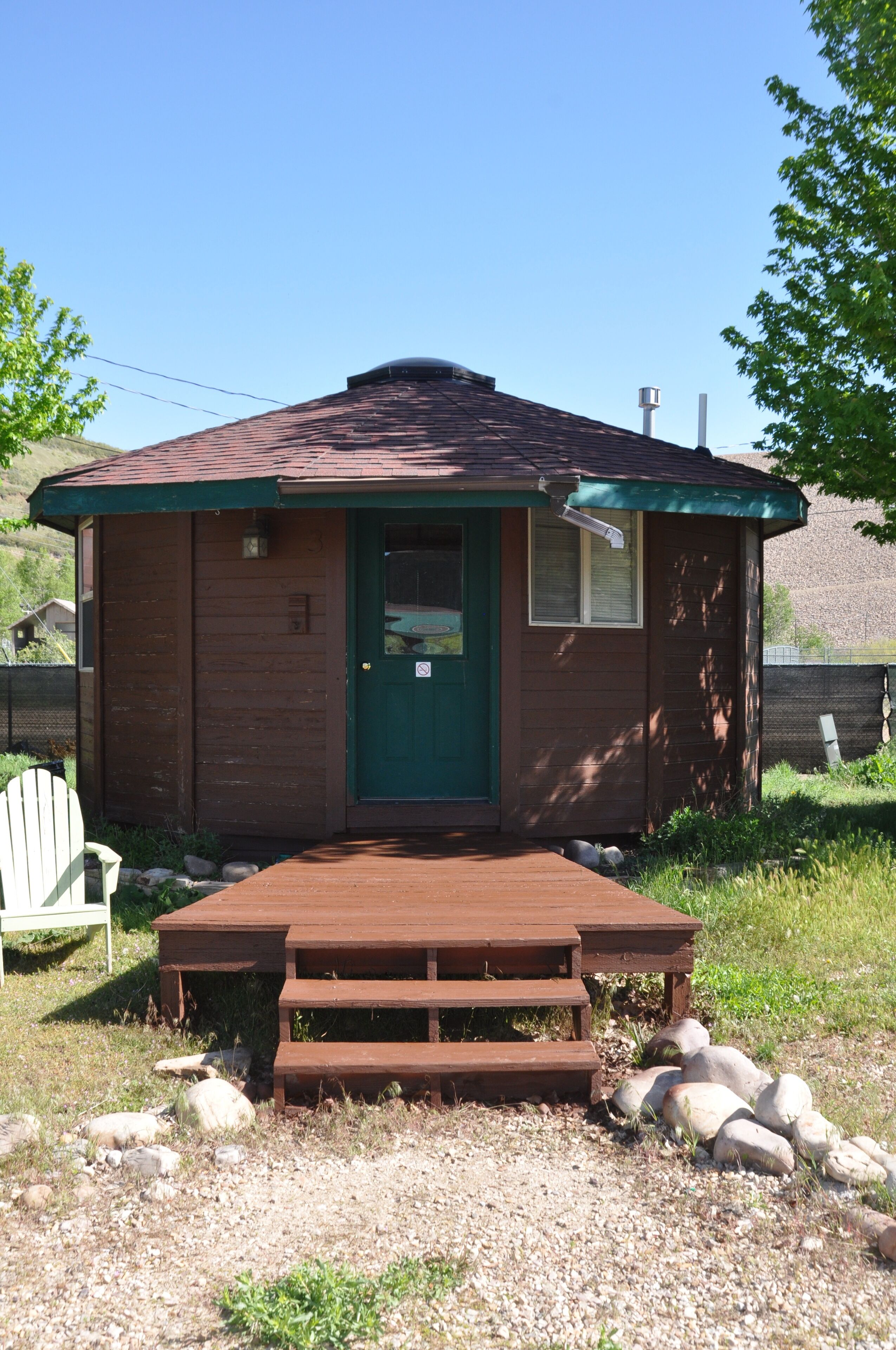 A small, single-story cabin with a green door and a small window, surrounded by a wooden deck and gravel ground.
