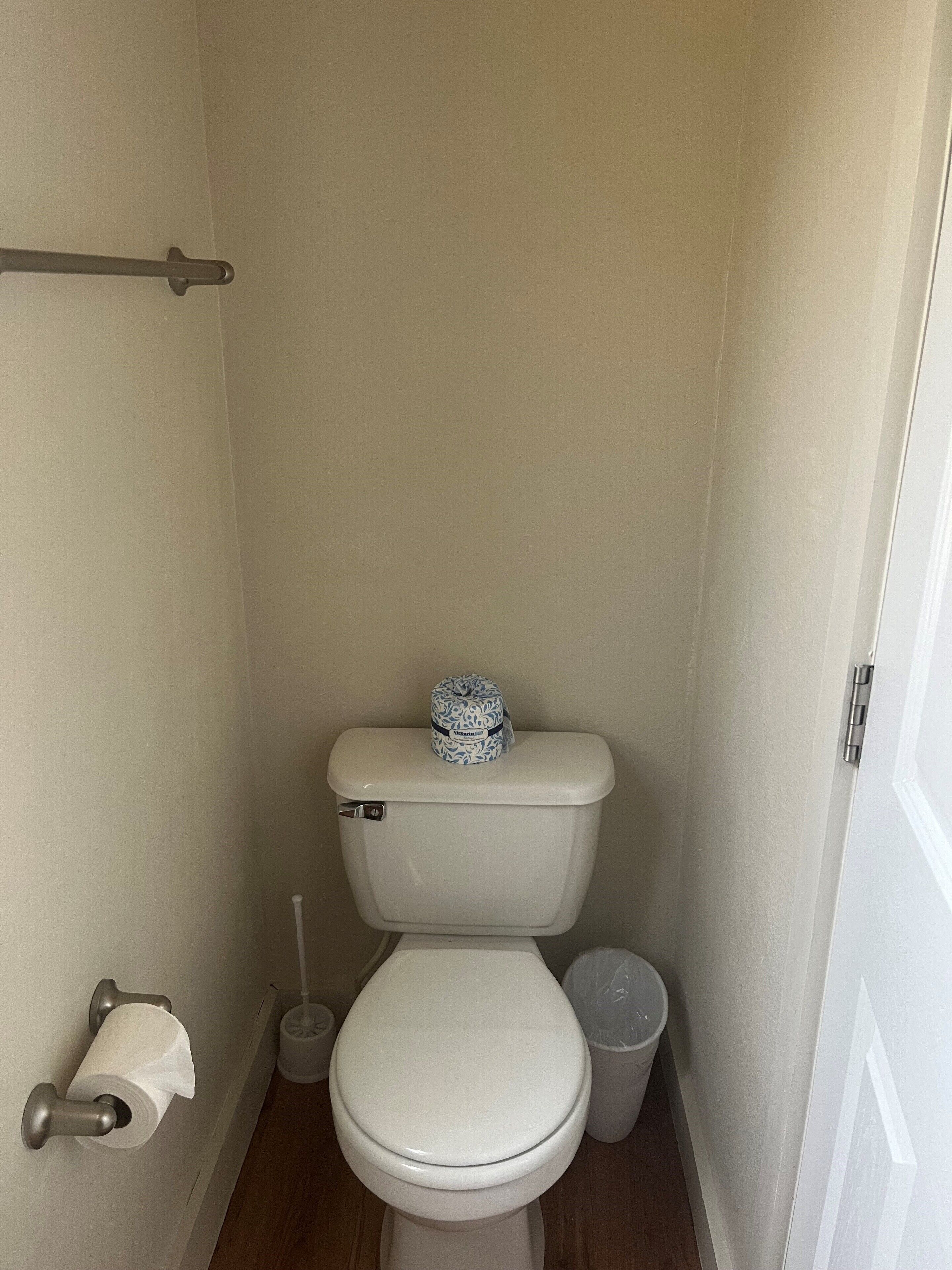 A small, clean bathroom with a toilet, a roll of toilet paper, a trash can, and a toilet brush.