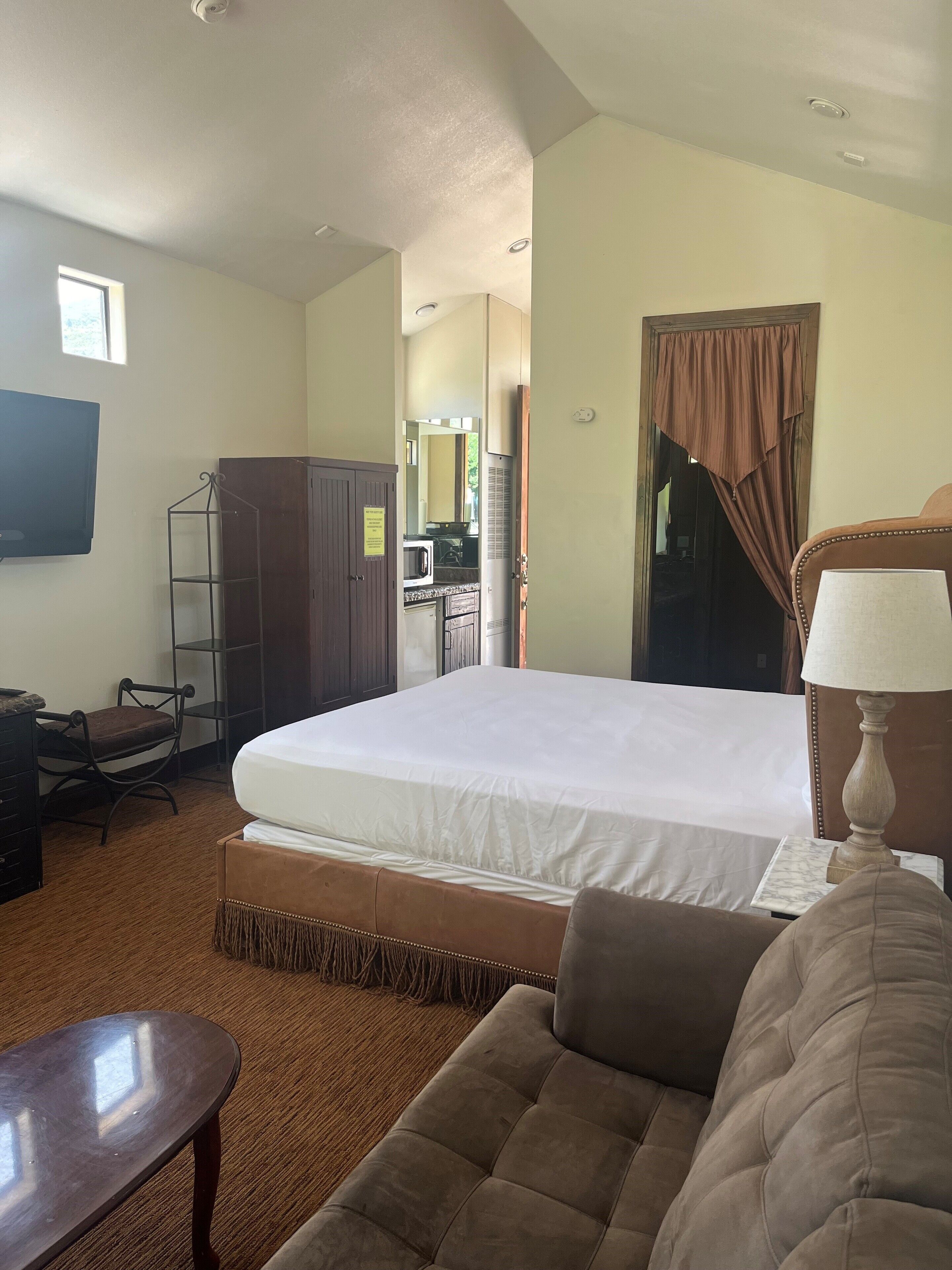 A hotel room with a bed, a desk, a chair, a TV, and a kitchenette in the background.