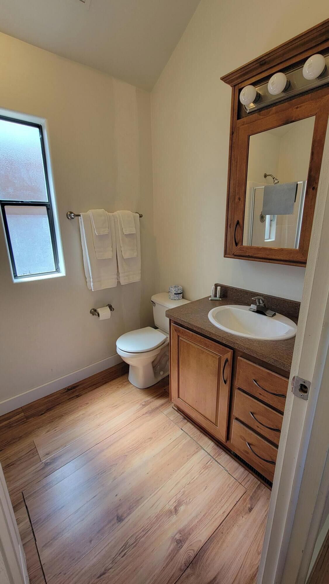 A bathroom with a wooden floor, a white toilet, a sink with a wooden cabinet, and a mirror above the sink.