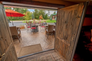 An open door leading to an outdoor patio with a table and chairs, a red umbrella, and a view of a road and trees.