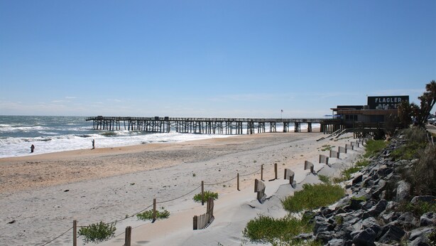 A beach with a wooden pier extending into the ocean, a building labeled 'FLAGLER', and a rocky shoreline with a fence. - Stunning Ocean Views, Sleeps 10, New Build, Beach! (Flagler Beach)