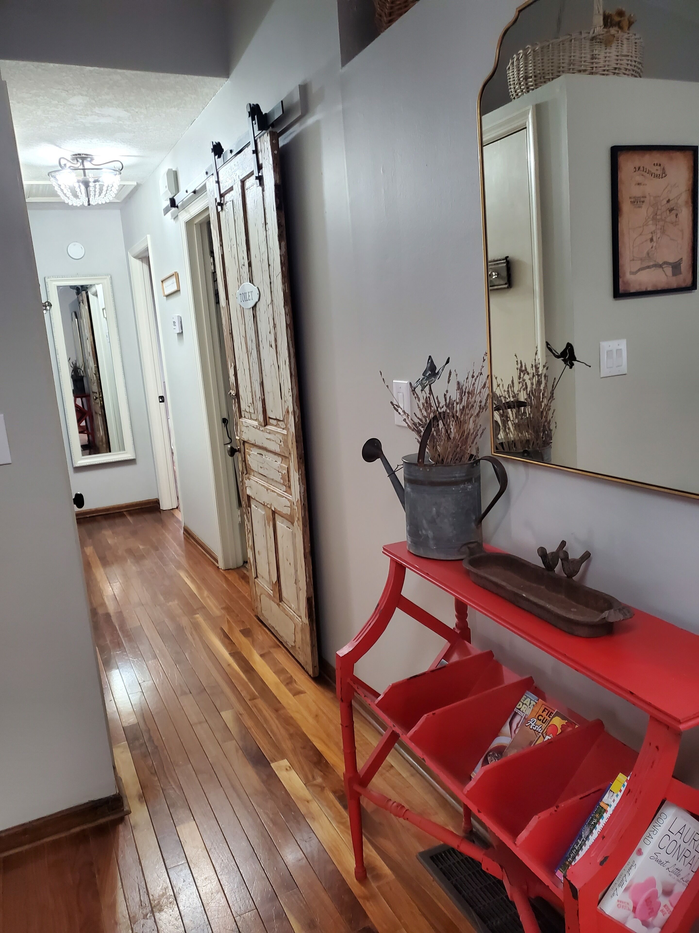 A hallway with wooden flooring, a red bench with magazines, a mirror, and a door labeled "TOILET".