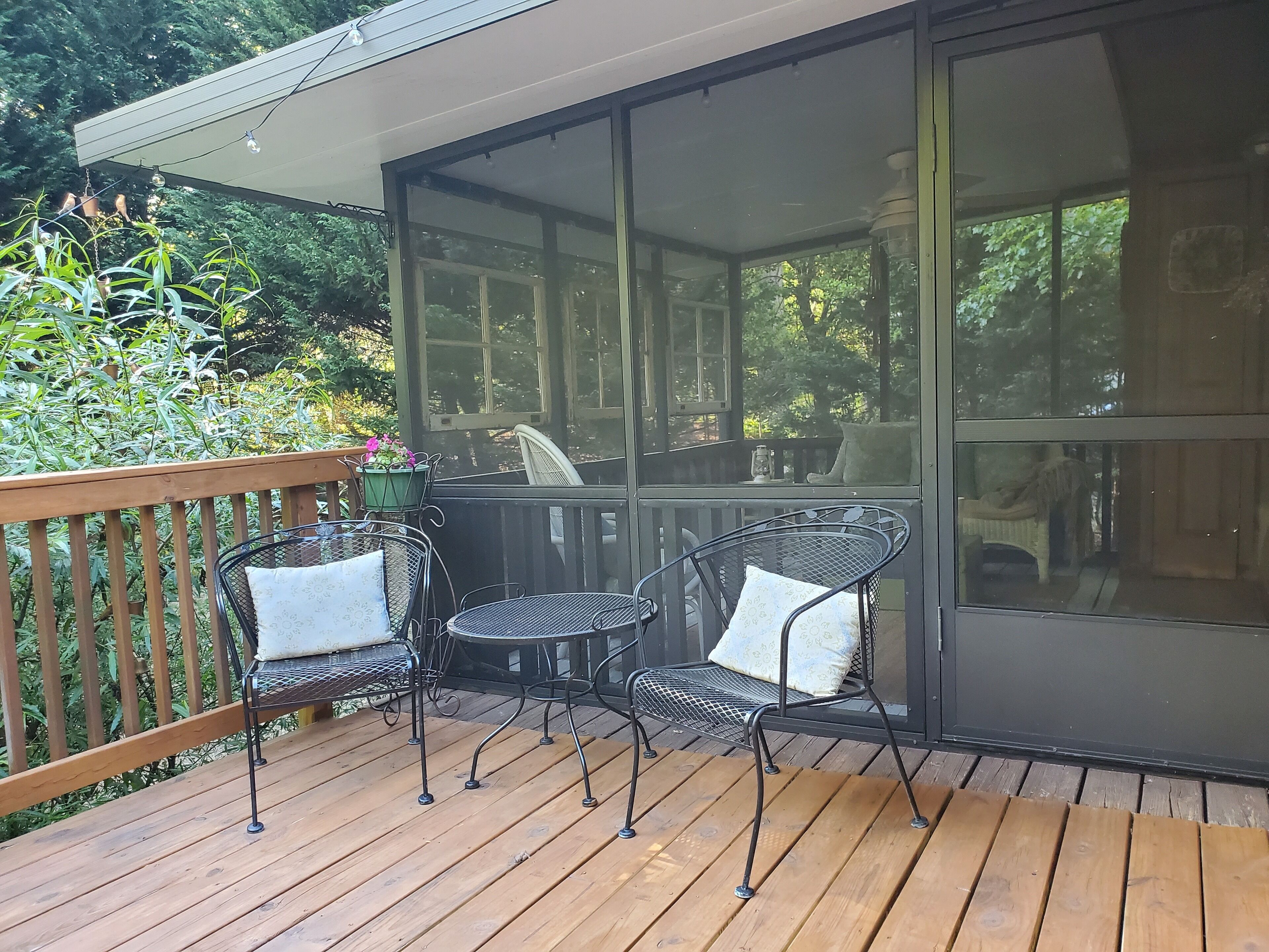 Une terrasse en bois avec deux chaises en métal et une table, une porte vitrée et une vue sur la verdure.