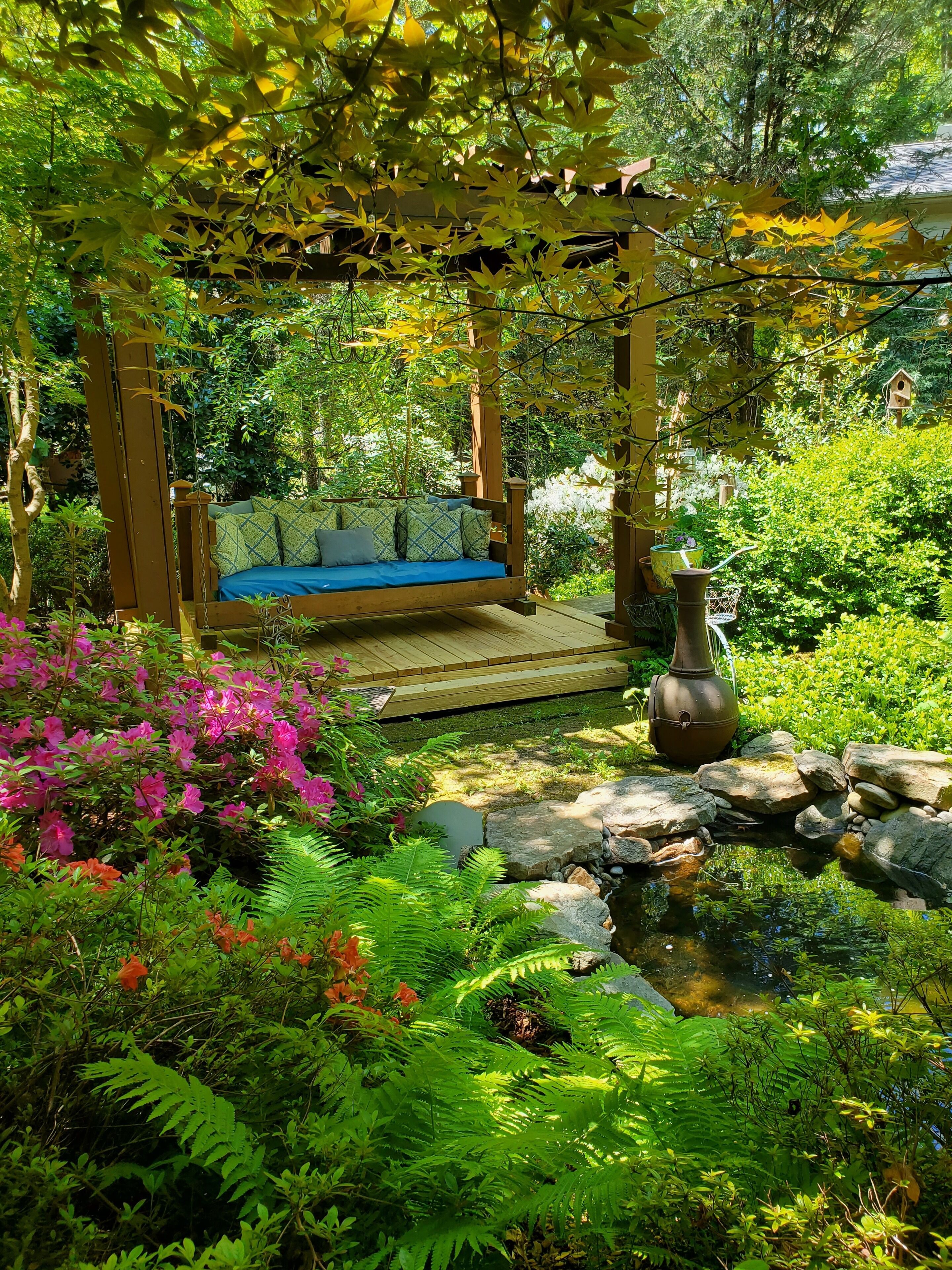 A wooden swing under a pergola, surrounded by lush greenery and a small pond.