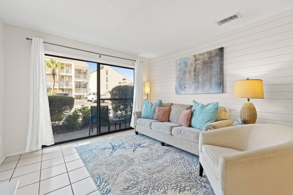 A modern living room with a sliding glass door leading to a balcony, a beige sofa with patterned cushions, a floor lamp, and a piece of abstract art on the wall.