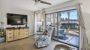 A living room with a flat-screen TV, wooden cabinet, patterned armchair, and a view of the outdoors through large sliding glass doors. - Endless Summer By Book That Condo (Panama City Beach)