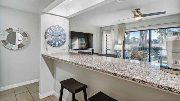 A modern kitchen with a granite countertop, a large clock, a flat-screen TV, and a ceiling fan. - Endless Summer By Book That Condo (Panama City Beach)