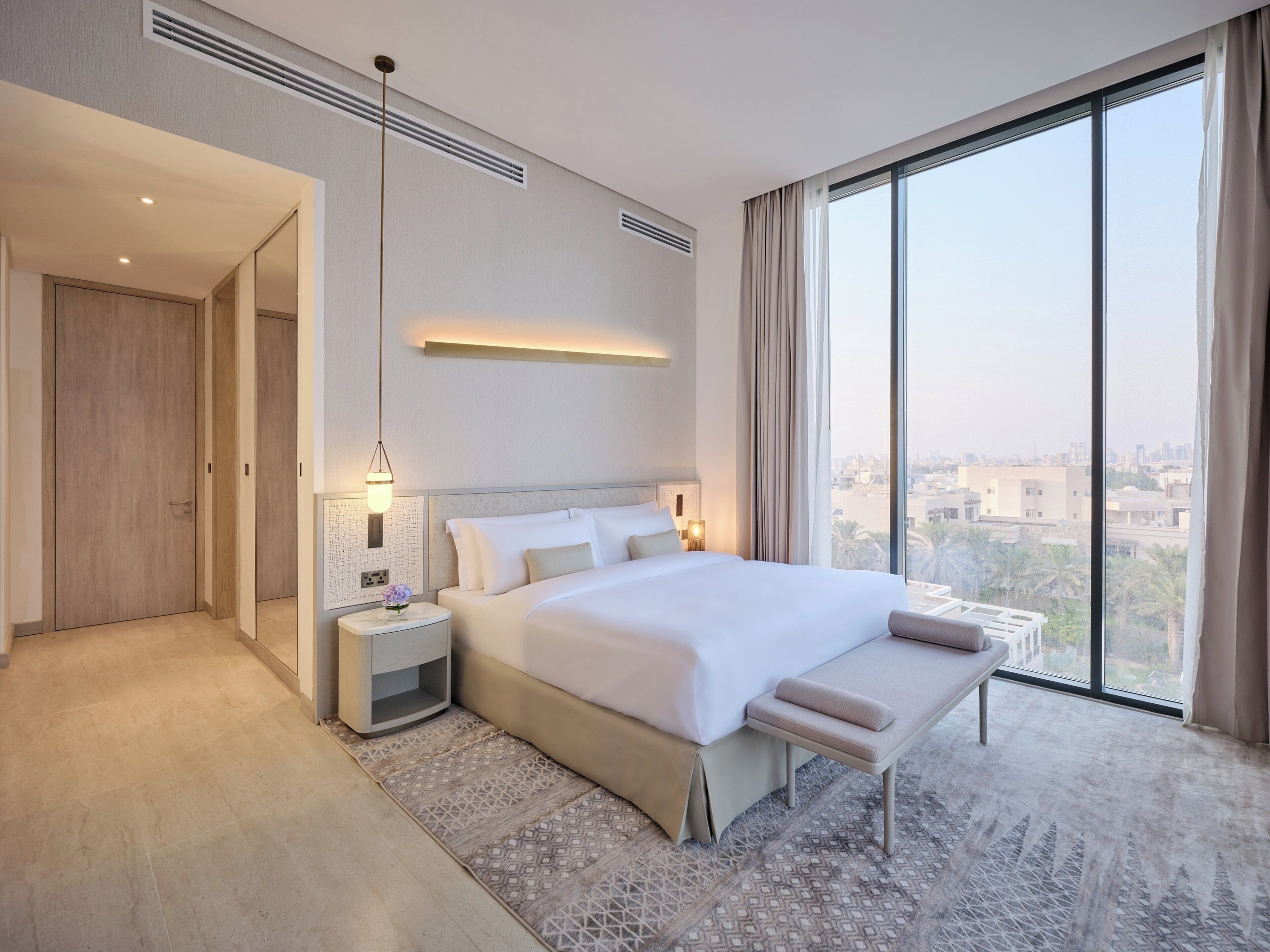 Una habitación de hotel moderna con una cama grande, mesitas de noche, un banco y vistas a la ciudad.