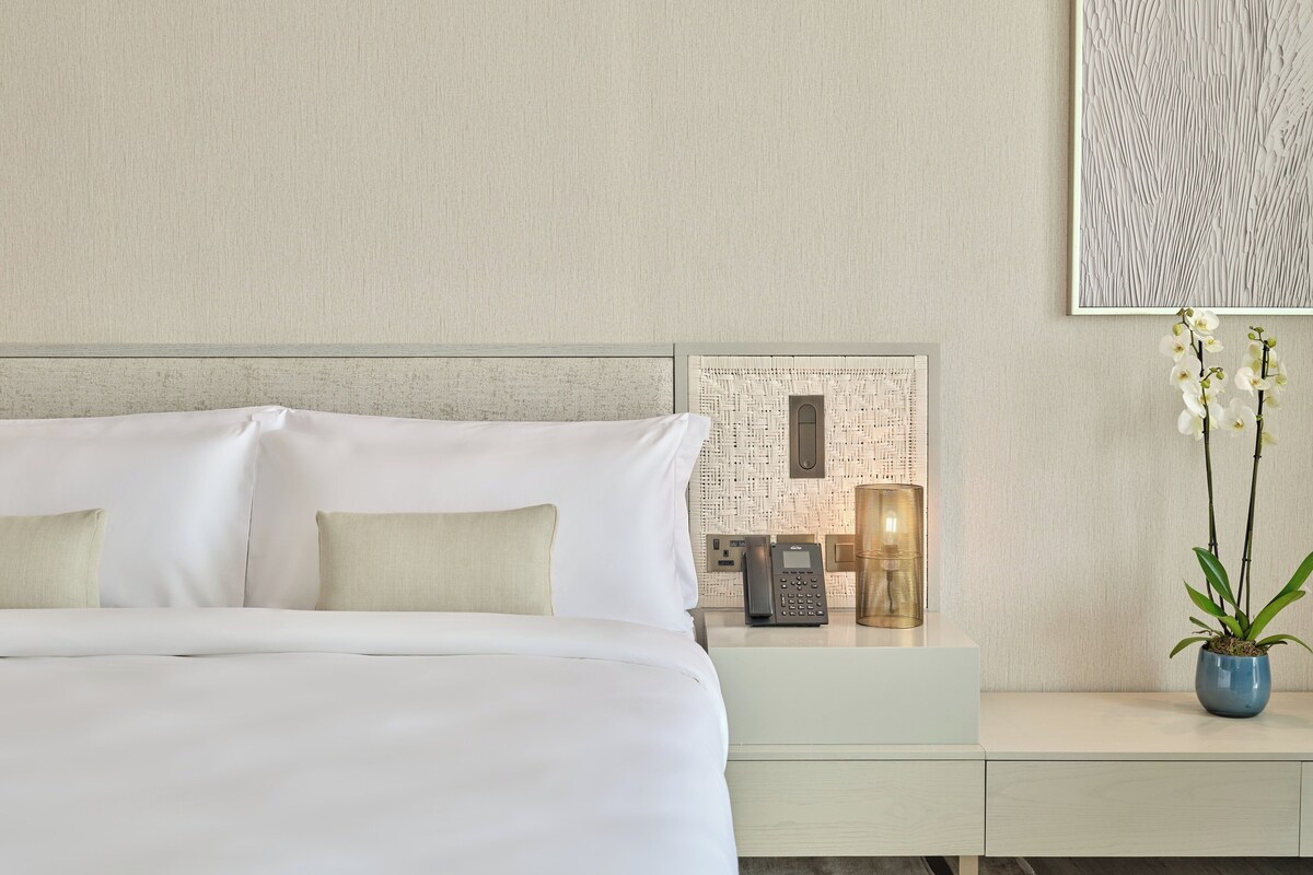 Een moderne hotelkamer met een groot bed, een nachtkastje met telefoon en lamp, en een kleine plant op een bijzettafeltje.