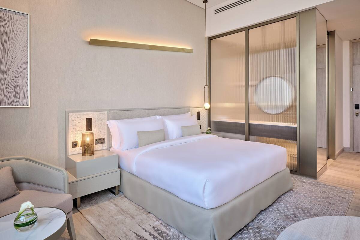 A modern hotel room with a large bed, bedside tables, a nightstand, a chair, and a small table with a vase.