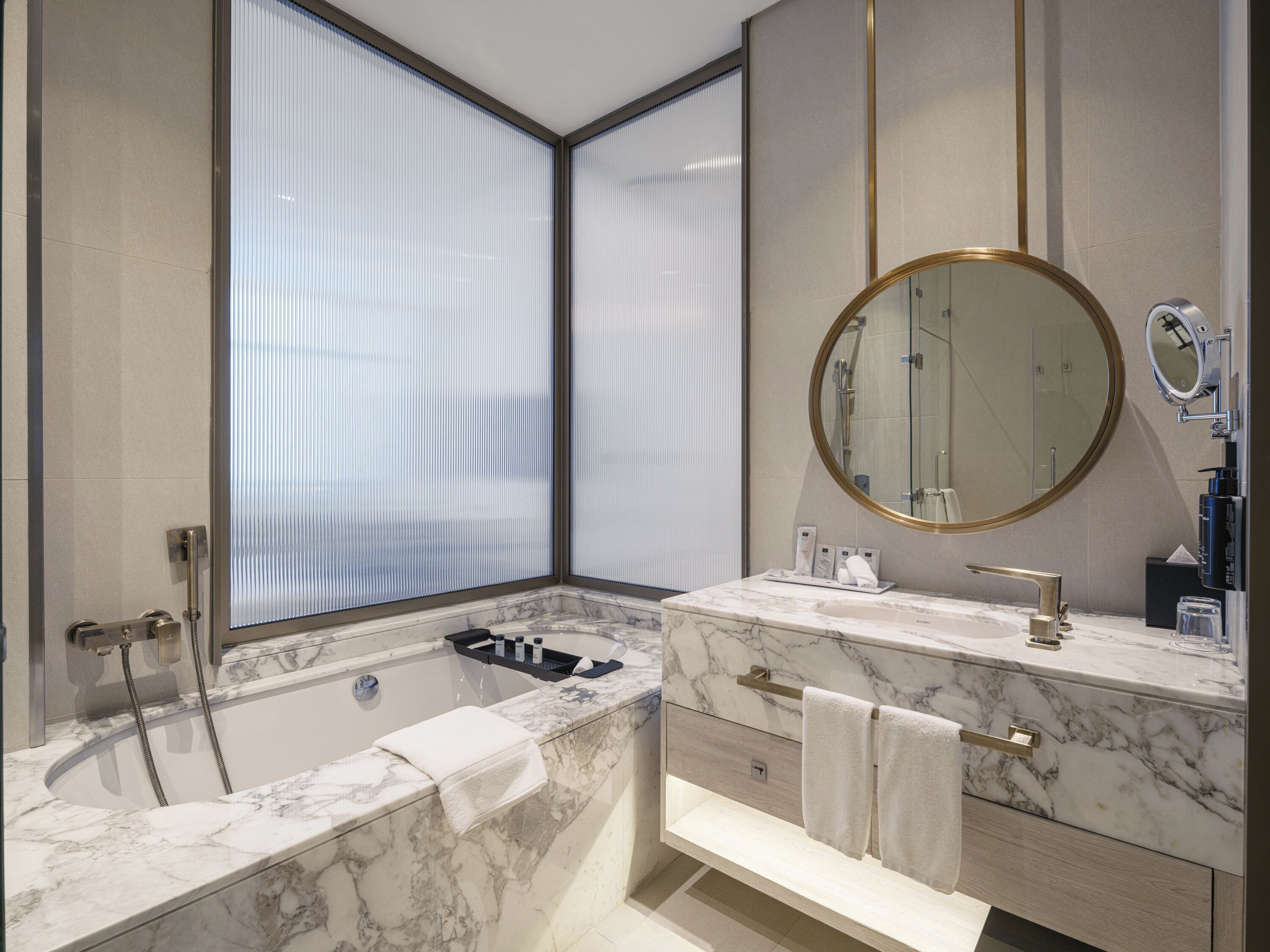 A modern bathroom with a bathtub, a vanity with a marble countertop, a round mirror, and a window with blinds.