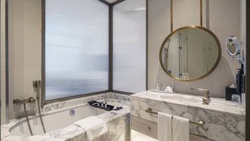 Un baño moderno con una bañera, un tocador con encimera de mármol, un espejo redondo y una ventana con persianas.