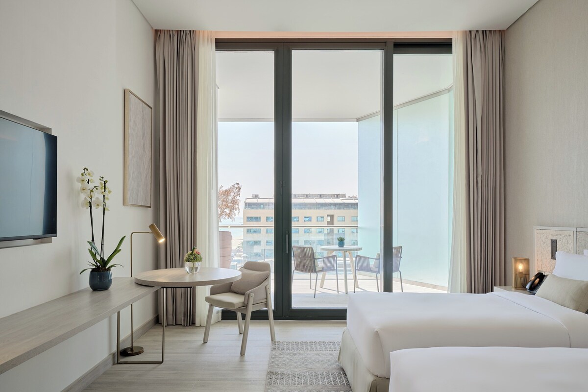 Een moderne hotelkamer met een groot bed, een bureau met een stoel, een kleine tafel met twee stoelen en een balkon met uitzicht op gebouwen.