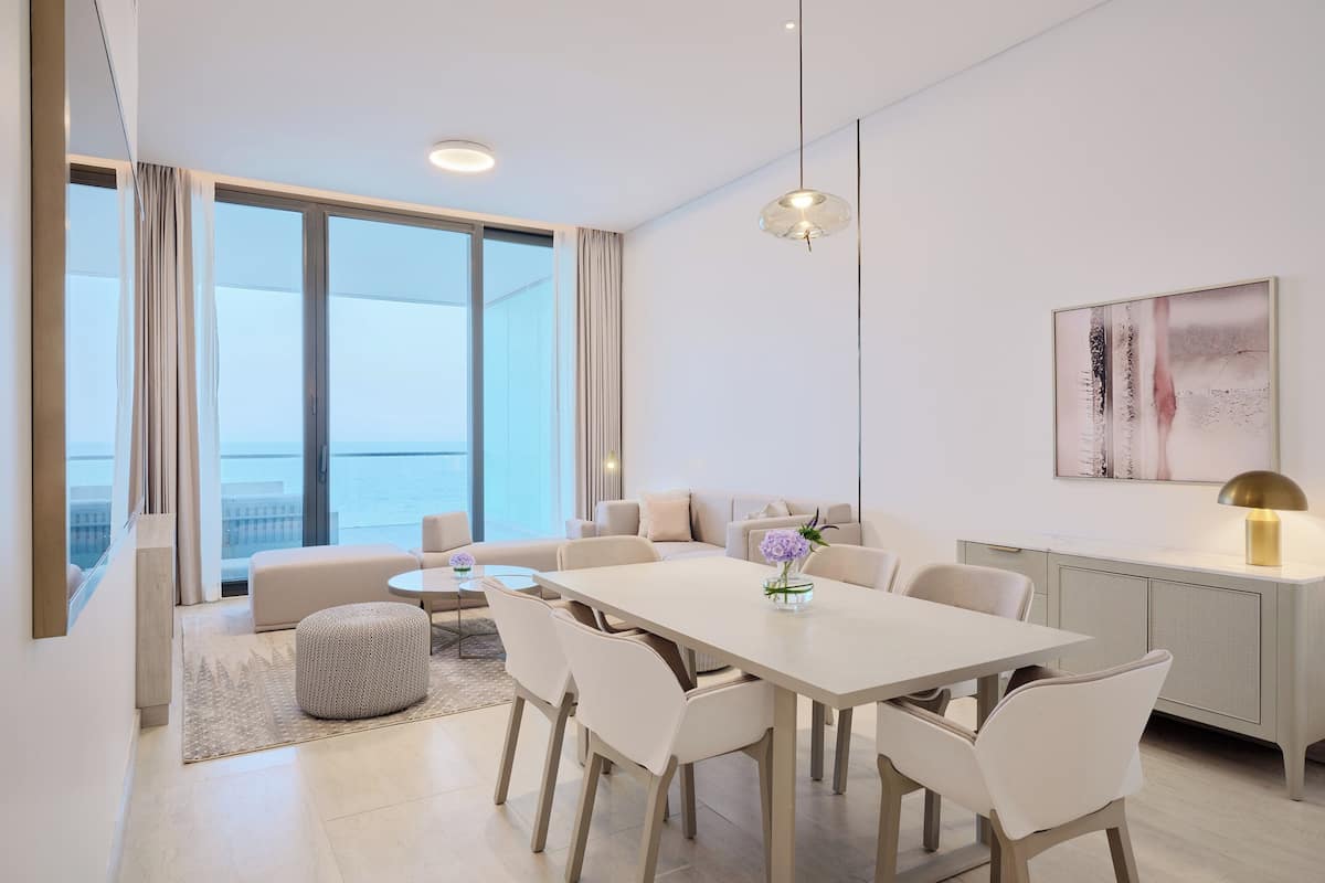 A modern hotel room with a large window offering a view of the sea, a dining area with a white table and chairs, and a minimalist decor.