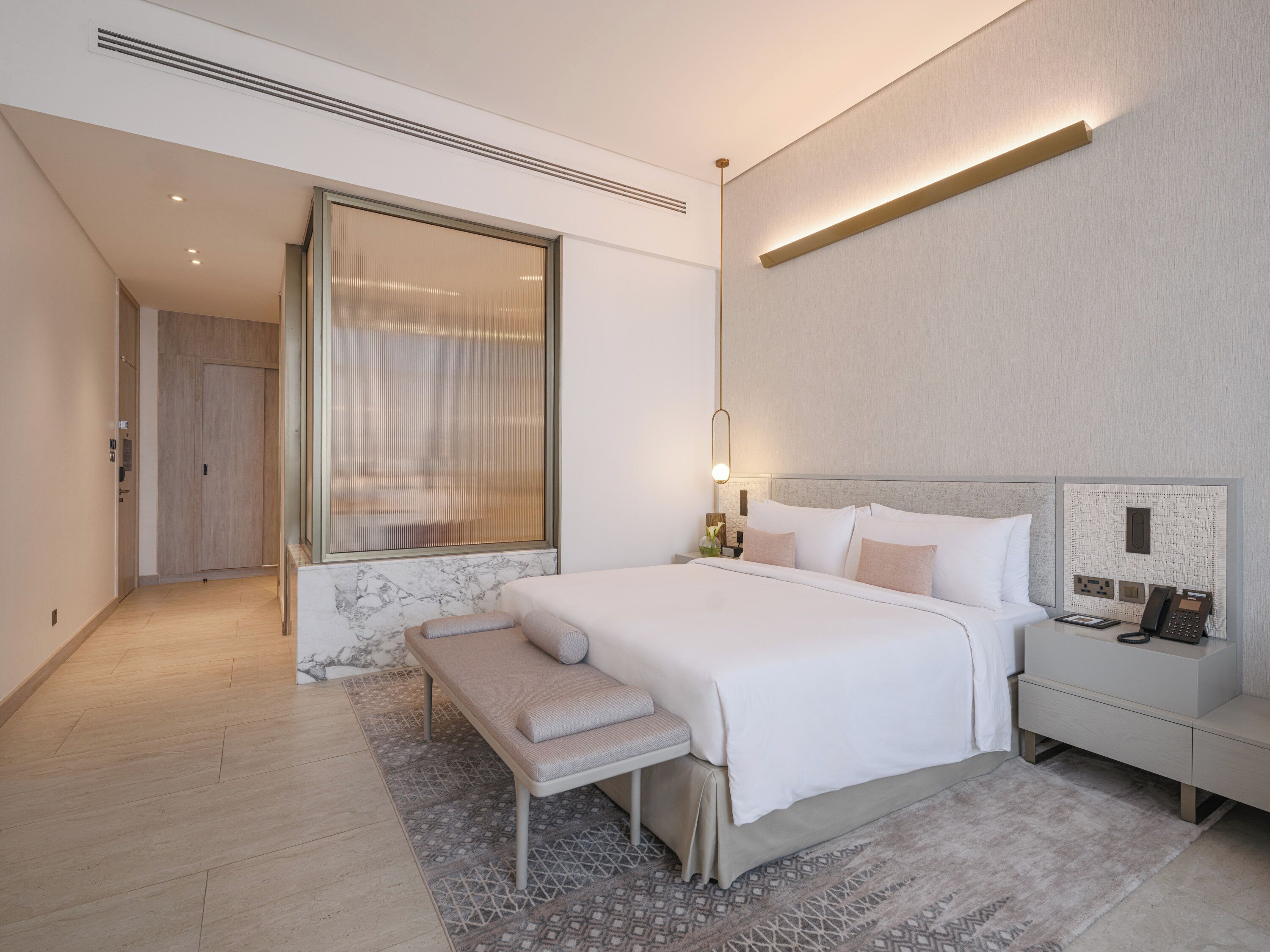 A modern hotel room with a large bed, bedside table, and a bench.