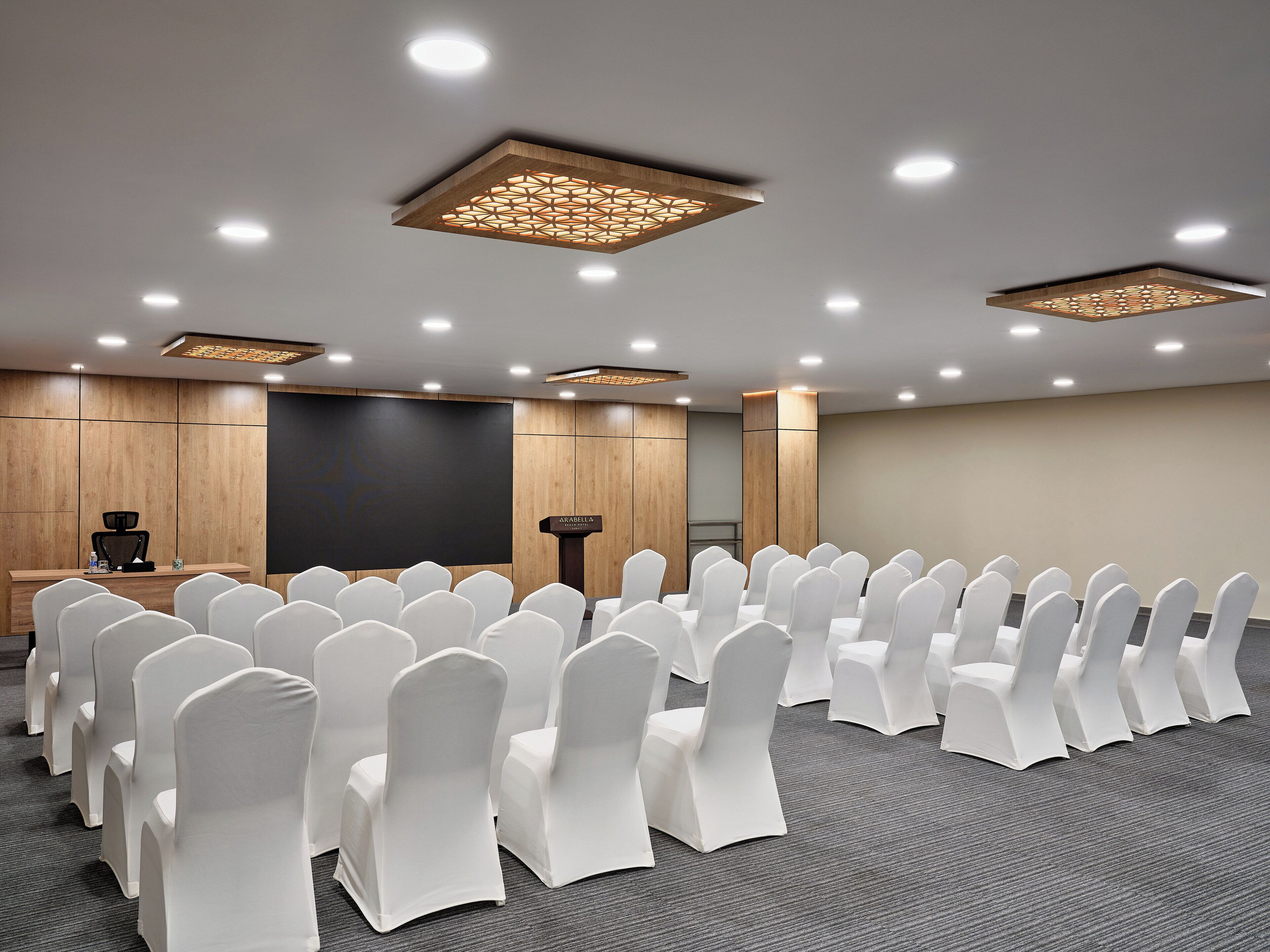 A conference room with rows of white chairs, a podium, and a large screen.
