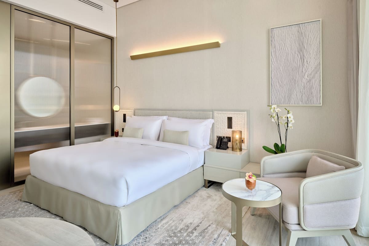 A modern hotel room with a large bed, bedside tables, a small round table, a chair, and a decorative wall piece.