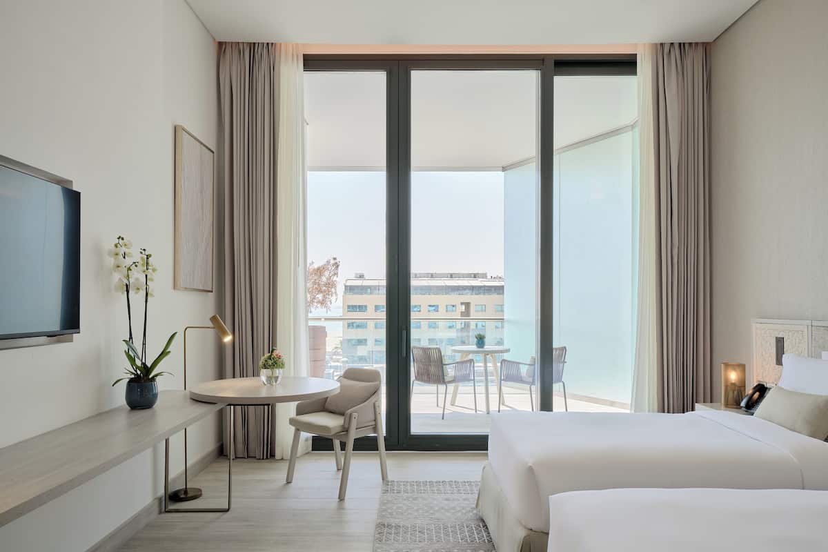 A modern hotel room with a large bed, a desk with a chair, a small table with two chairs, and a balcony with a view of buildings.