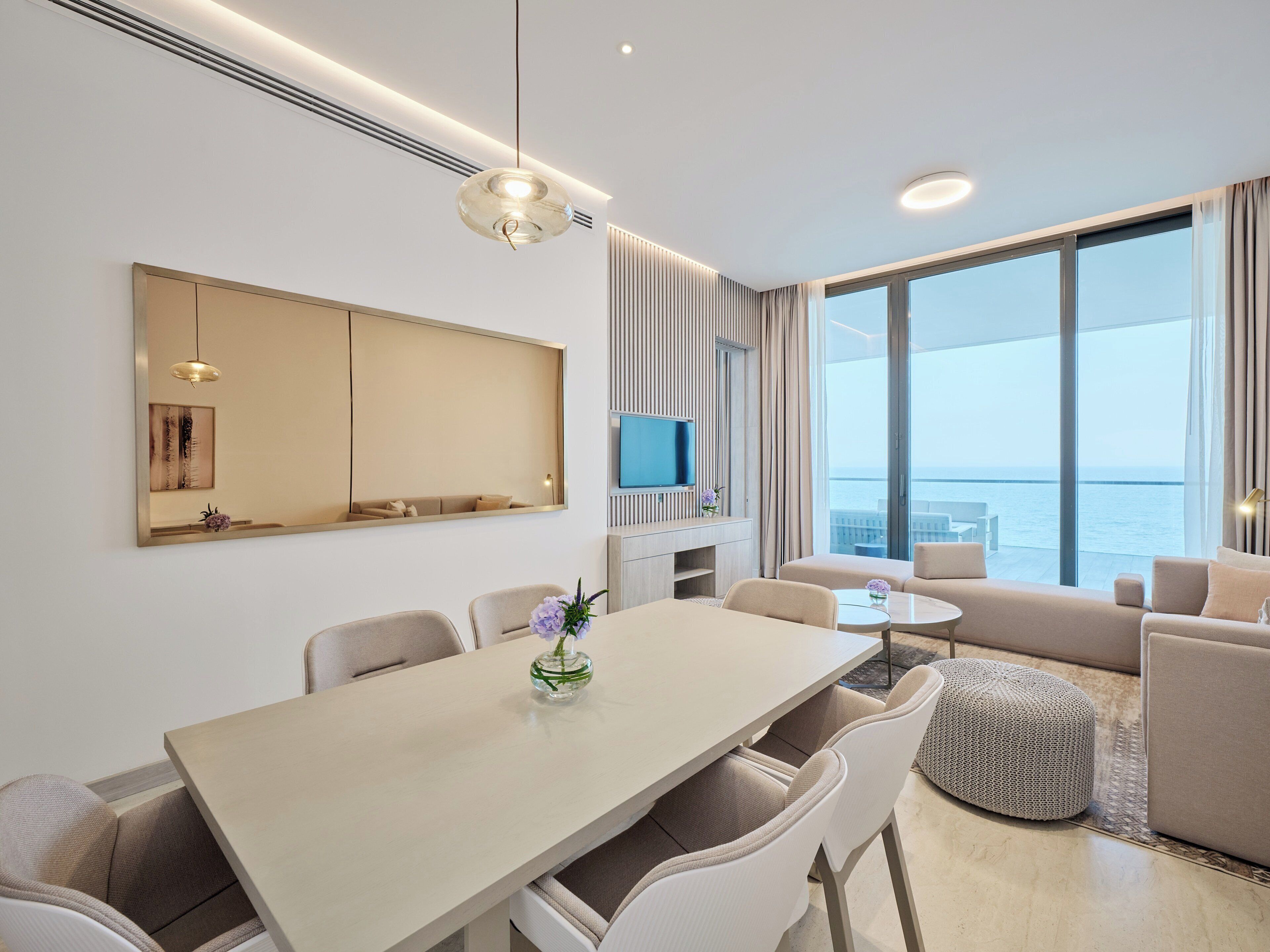 A modern hotel room with a dining area, a living space, and a large window offering a view of the sea.