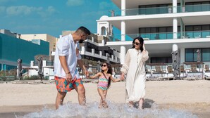 Een gezin wandelt over het strand, met een modern hotel op de achtergrond.