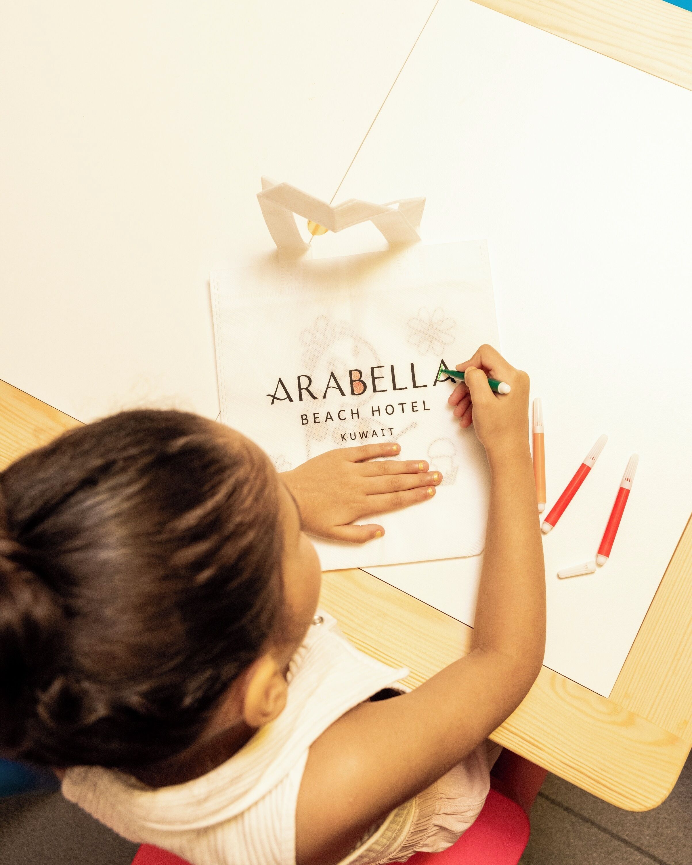 A child drawing on a paper with the text "ARABELLA BEACH HOTEL KUWAIT" visible.
