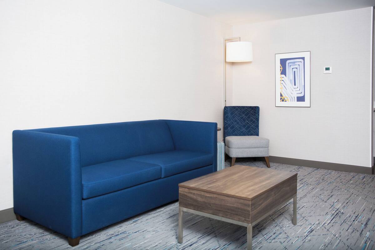 Een kamer met een blauwe bank, een houten salontafel, een grijze fauteuil, een vloerlamp en een lijst met abstracte kunst aan de muur.
