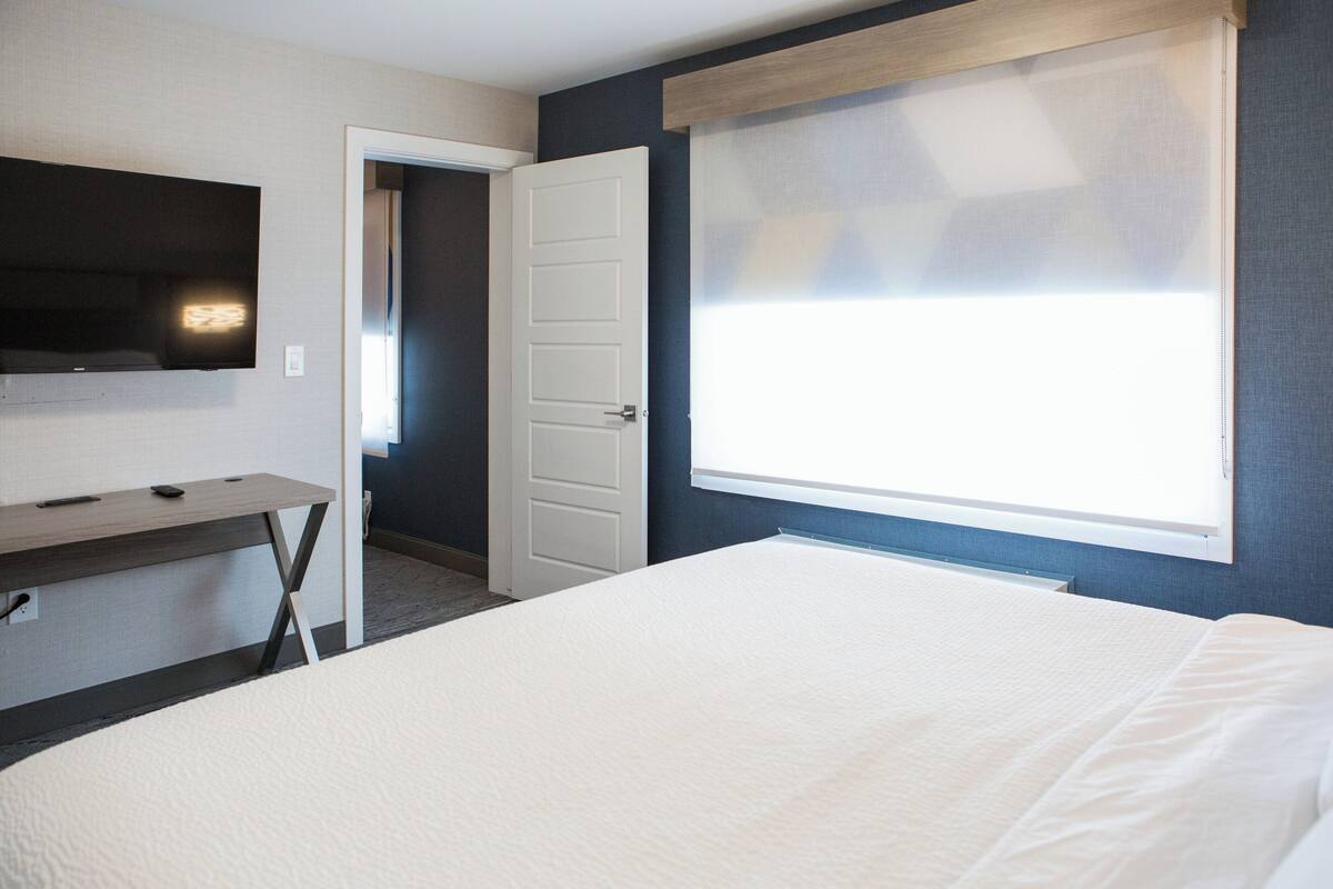 Een slaapkamer met een bed, een bureau, een televisie en een raam met zonwering.