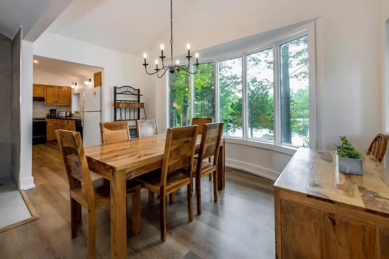 A spacious dining area with a wooden table and chairs, a chandelier, and large windows offering a view of the outdoors.