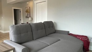 A grey sectional sofa with a wooden side table in a room with light wooden flooring and a white door in the background. - Full Comfy&Private Suite with a Yard for Your Getaway in Bear Mountain, Victoria (Victoria)