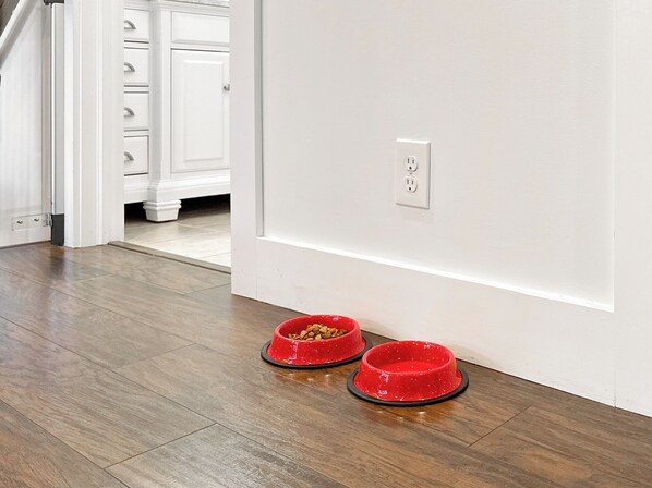 Una cocina moderna y limpia, con piso de madera y armarios blancos. Hay dos platos rojos para mascotas en el suelo.